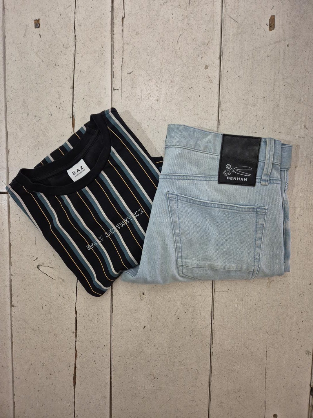 Flatlay met een zwart gestreept DAZ-shirt naast een lichte Denham-denim op een licht houten ondergrond.